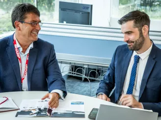 Two business men sat having conversation in a meeting room with a paper brochure and laptops